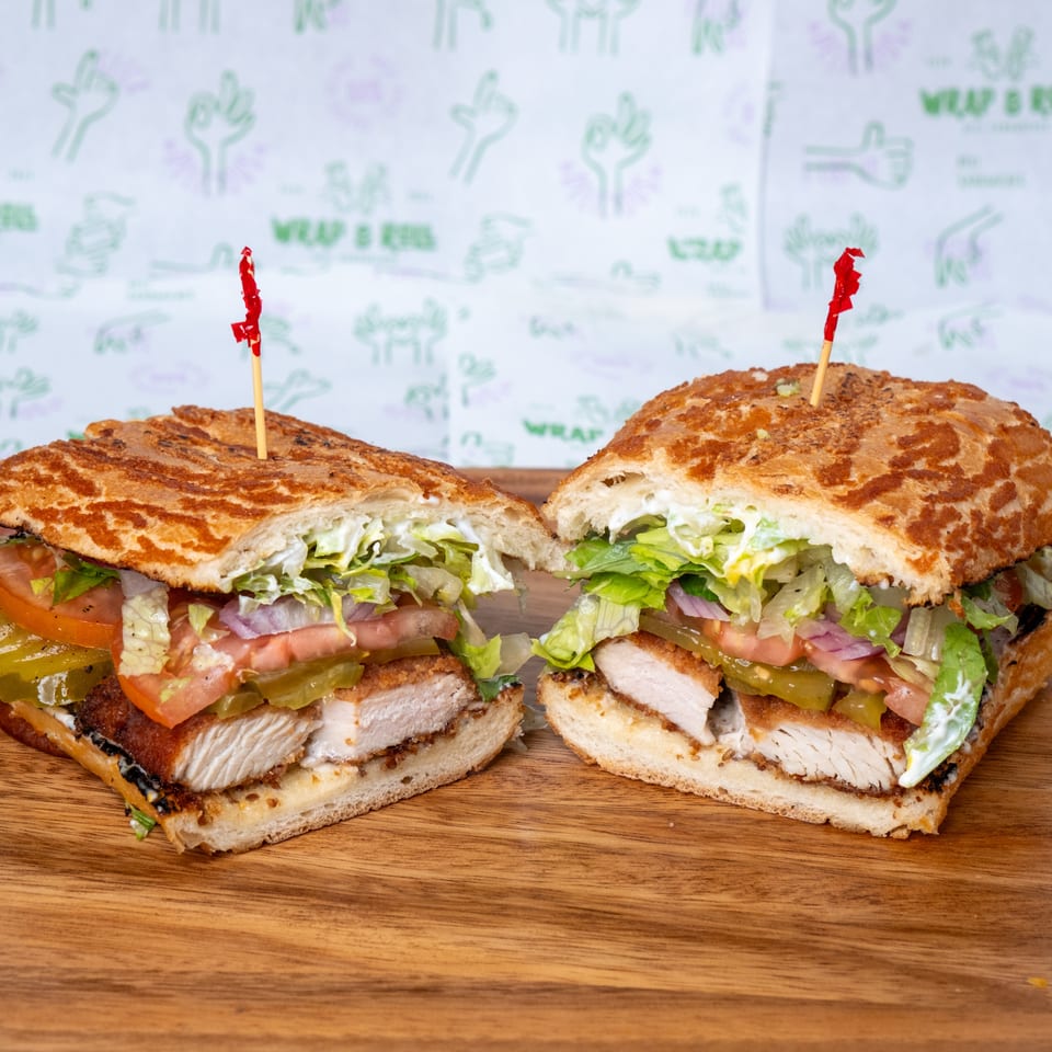 Two loaded chicken sandwiches with toasted bread, lettuce, tomatoes, pickles, onions, and creamy dressing on a wooden table.