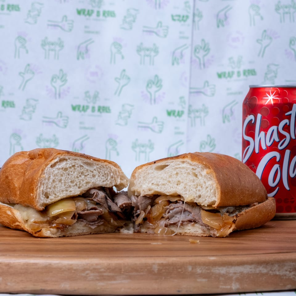 Sliced roast beef and melted cheese sandwich with caramelized onions on a wooden board beside a red Shasta soda can.