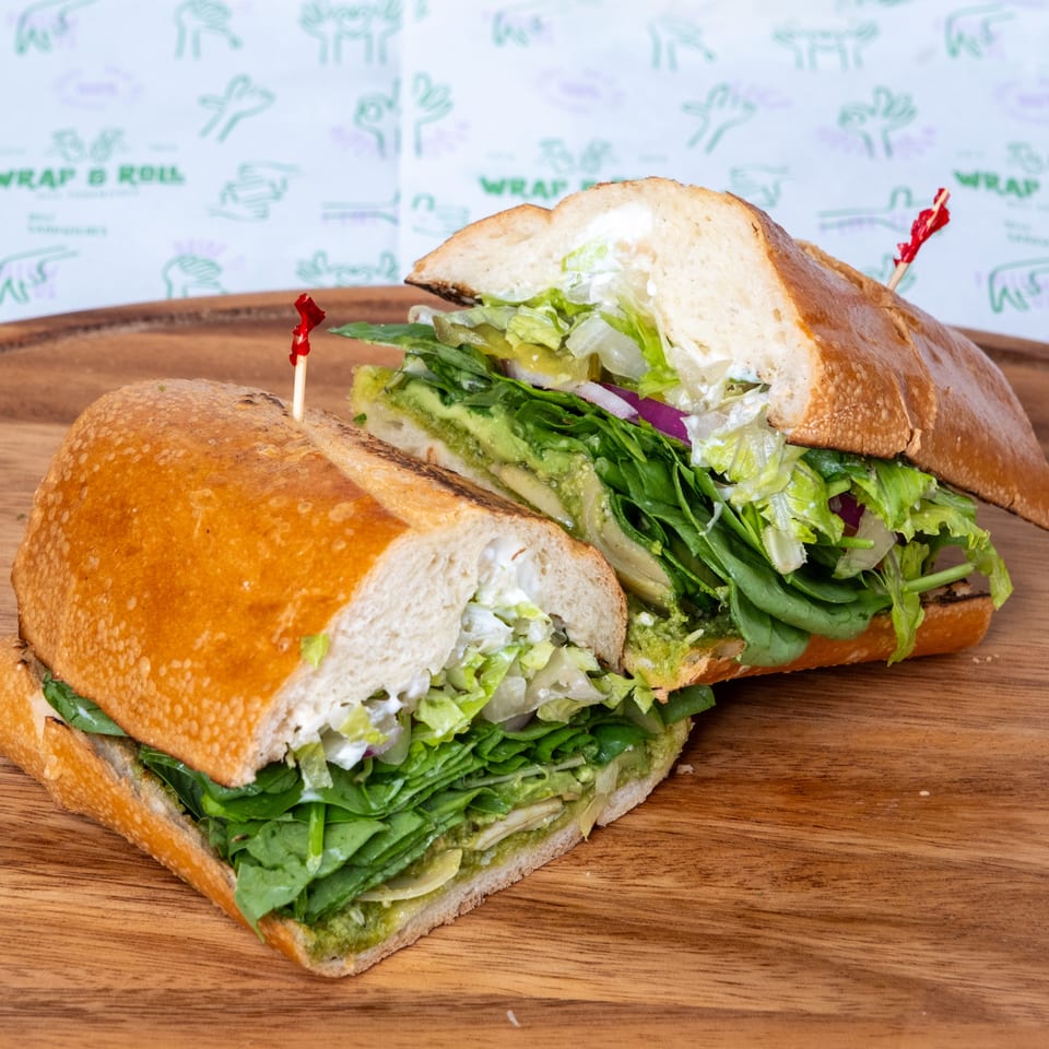 Two toasted vegetable sandwiches layered with lettuce, spinach, avocado spread, pickles, onions, and creamy dressing on a wooden table.
