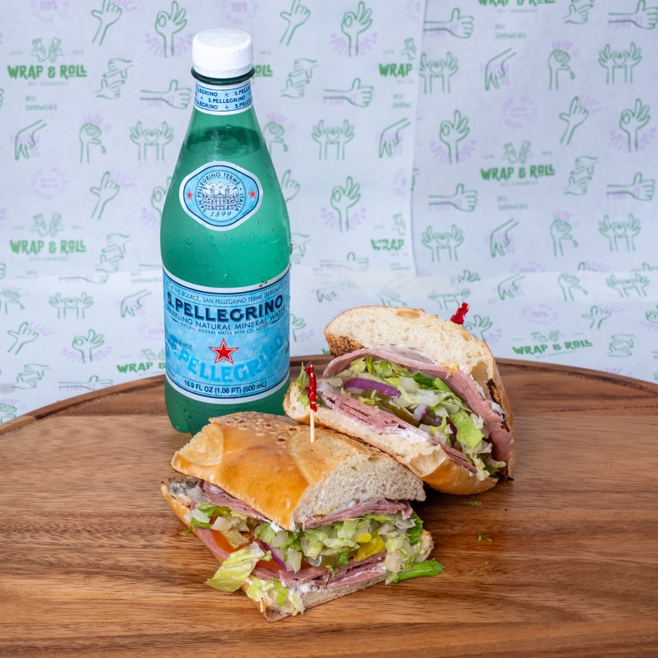 Two loaded deli sandwiches with lettuce and vegetables sit beside a chilled San Pellegrino bottle on a wooden serving board.