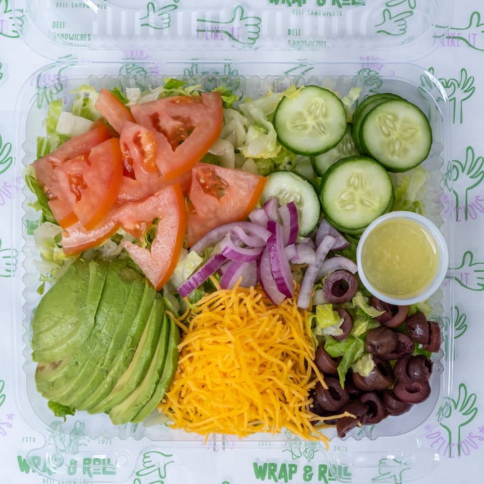 Open container of chopped salad with lettuce, tomatoes, cucumbers, avocado, red onion, cheddar cheese, olives, and dressing.