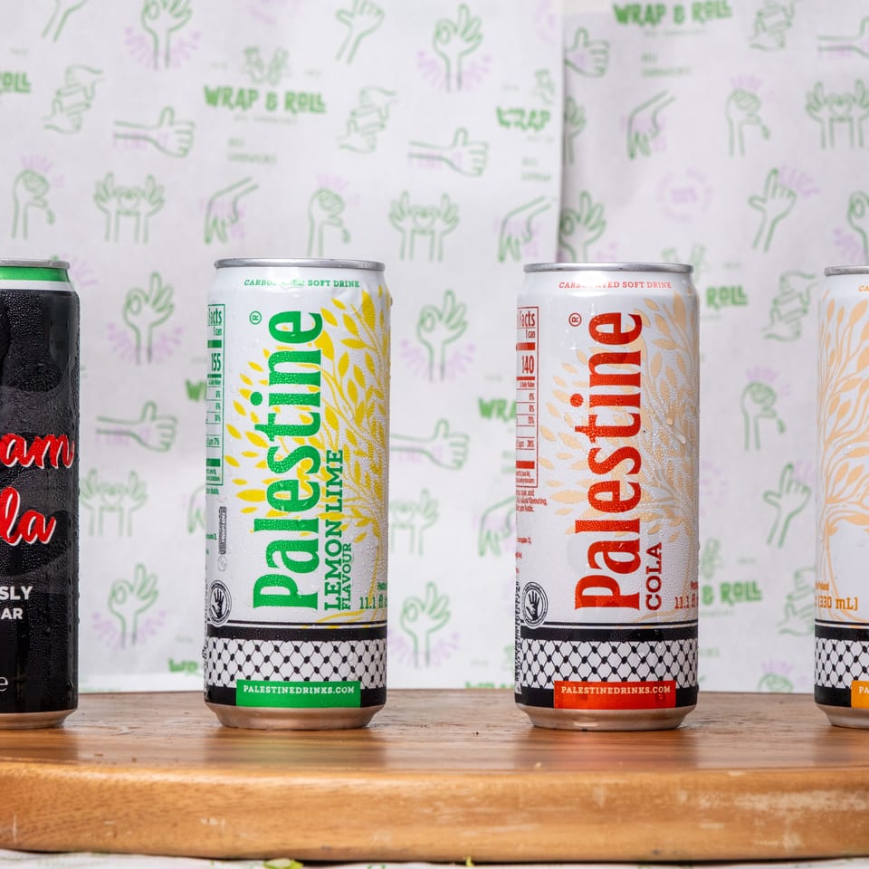 Four Palestinian soft drink cans, including cola, lemon-lime, cola, and orange flavors, displayed on a wooden counter.
